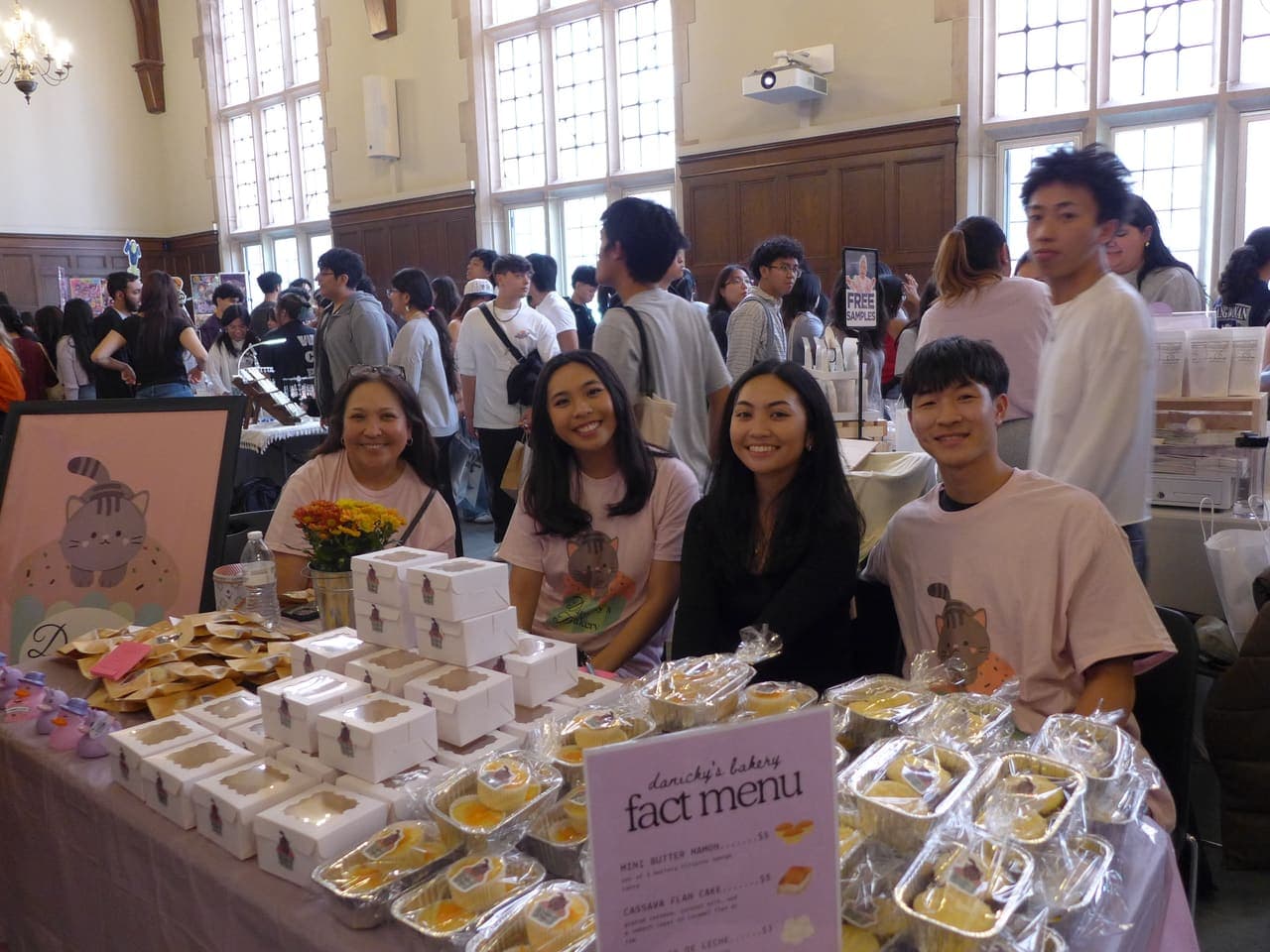 Volunteers behind a bakery table at FACT's Palengke market, smiling with trays of Filipino pastries
